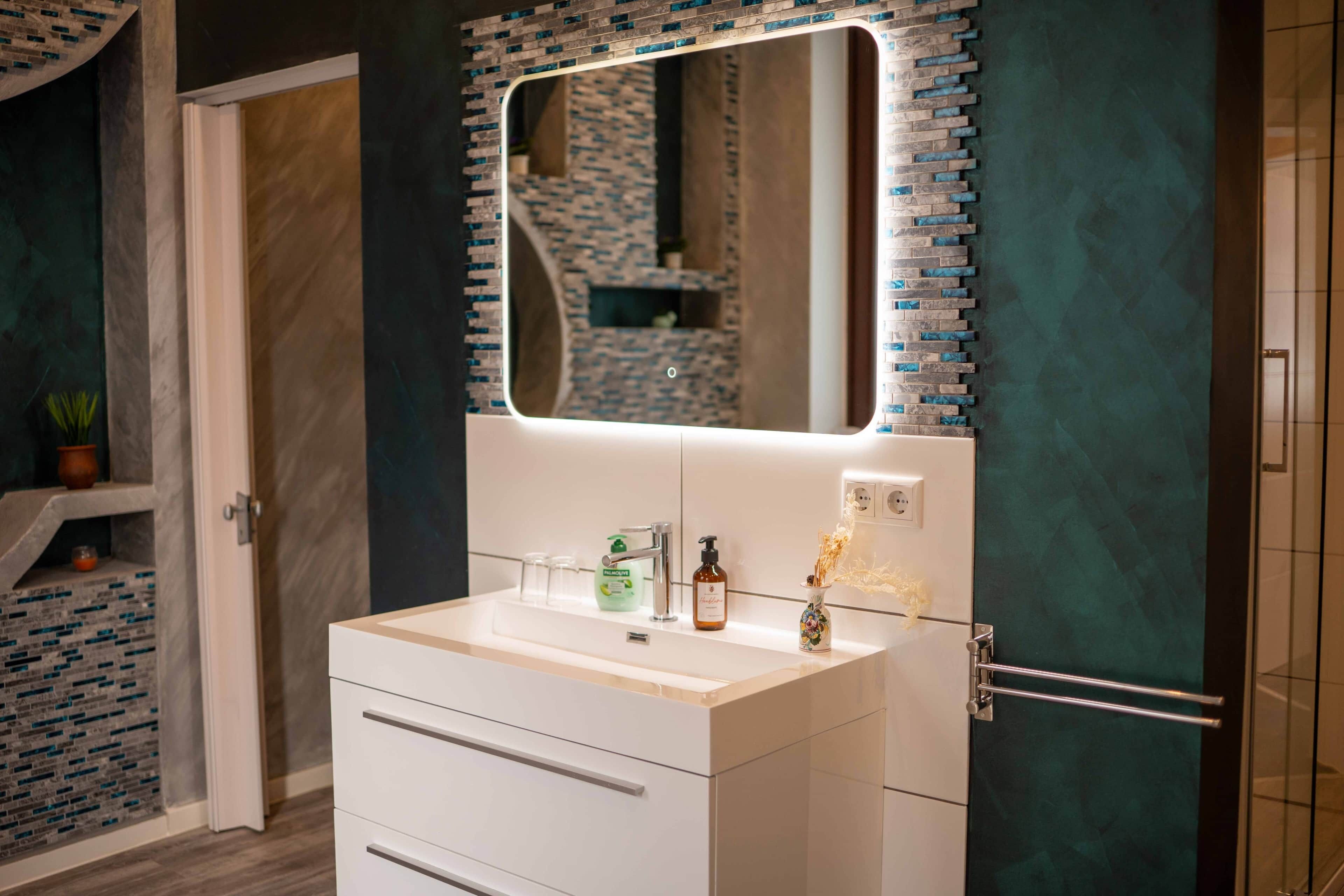 A bathroom with a white vanity, a mirror with a light, and a tiled backsplash.