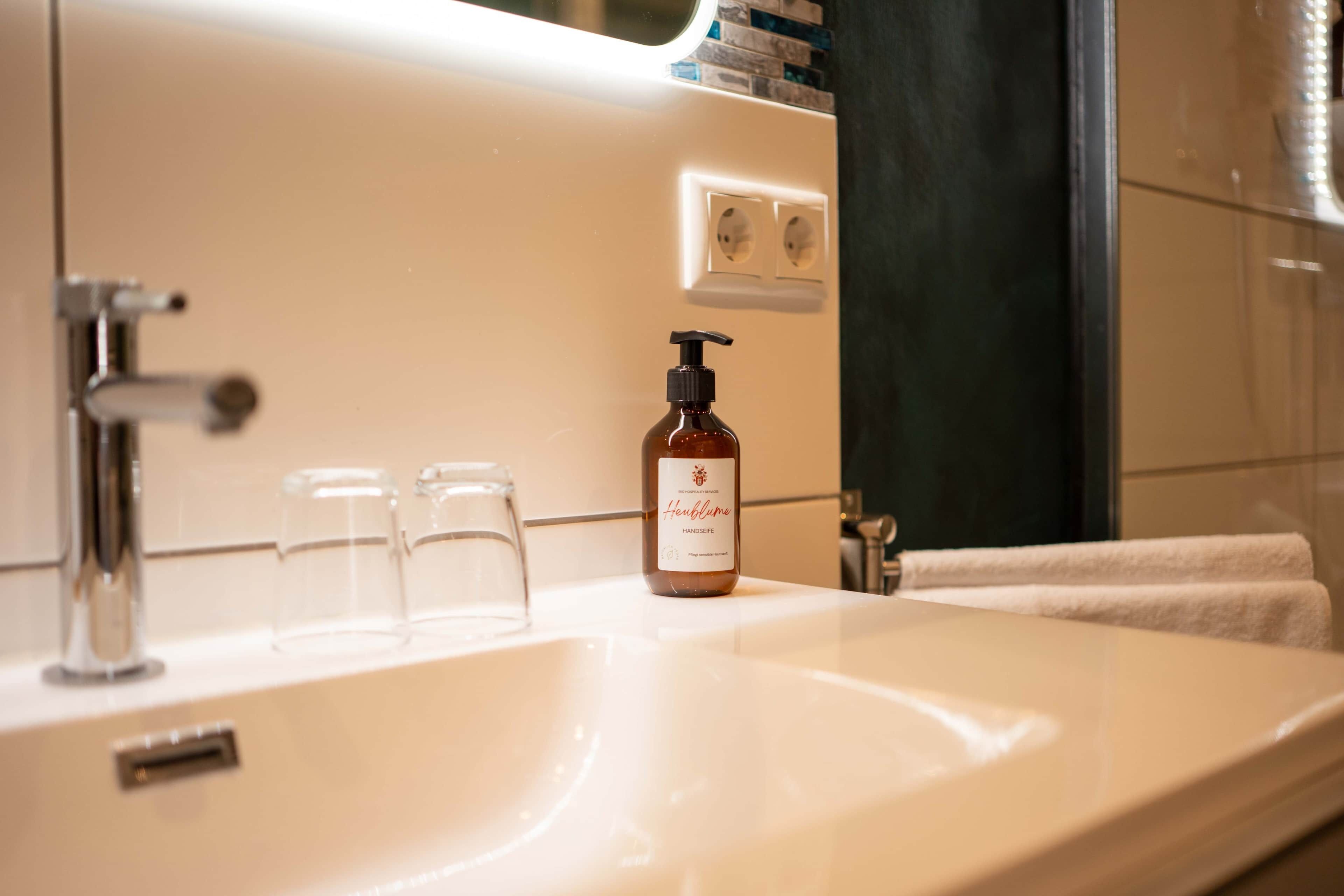Un baño con un lavamanos blanco, una botella de jabón de manos y un espejo.