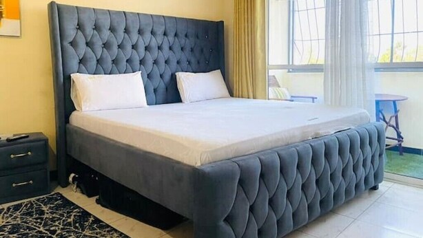 A bedroom with a tufted headboard, a bed with white linens, a small bedside table, and a view of the outdoors through a window. - A clean studio Apartment in Nyali, Mombasa. (Mombasa)