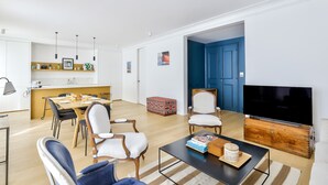 A modern living room with a dining area, a TV, and a kitchen in the background. - Stylish 2BR in Private Mansion at Rue de Passy (Paris)