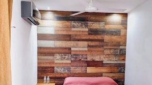 A bedroom with a wooden wall panel, a bed with a red cover, a nightstand, a ceiling fan, and an air conditioning unit. - Departamentos Túumben Ché (Valladolid)