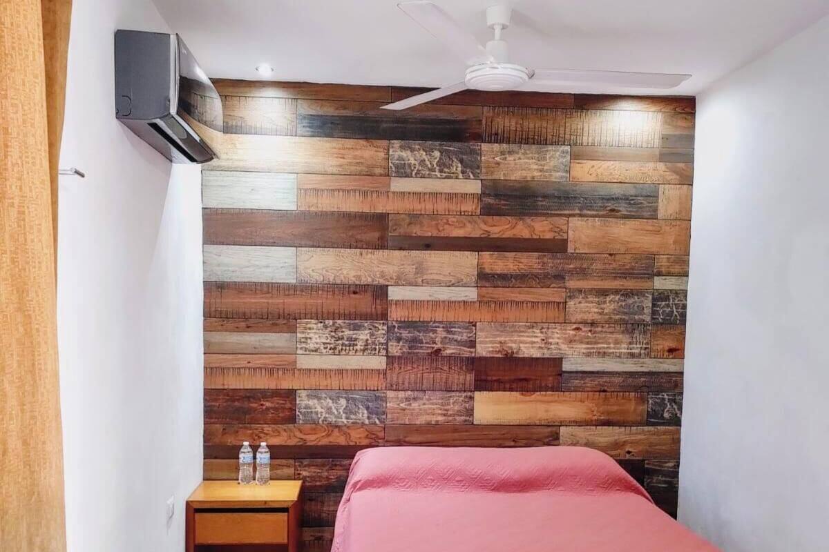 A bedroom with a wooden wall panel, a bed with a red cover, a nightstand, a ceiling fan, and an air conditioning unit.