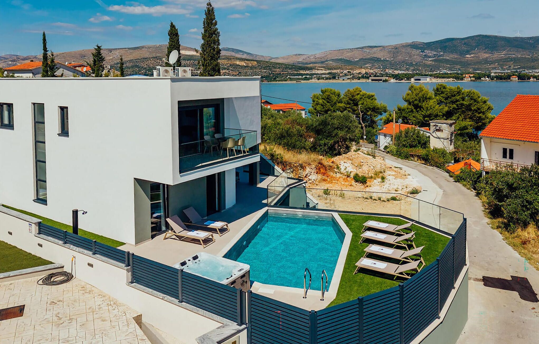 Un edificio moderno de dos pisos con piscina en la azotea, asientos exteriores y vistas a un lago y montañas.