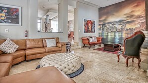 Un vaste salon agrémenté d’un canapé en cuir marron, d’un pouf rond, d’un fauteuil en bois et d’une fresque murale représentant un paysage urbain.