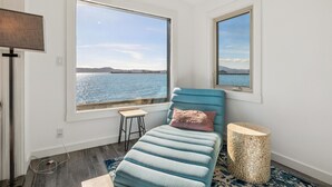 Ein Zimmer mit einem Bett, einem kleinen Hocker, einem runden Holztisch und großen Fenstern mit Blick auf das Wasser.