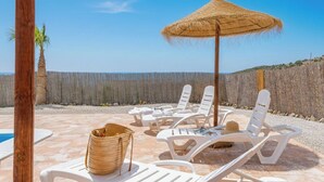 Ein Strandloungebereich mit weißen Liegestühlen, einem Strohdachschirm und einem Korbwannenstuhl.