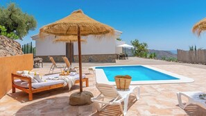 Un espace piscine avec des chaises longues, un parasol en chaume et un bâtiment en pierre.