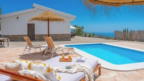 Un espace aménagé au bord de la piscine, avec des chaises longues, une table sur laquelle est posé un plateau de boissons et un parasol en chaume.