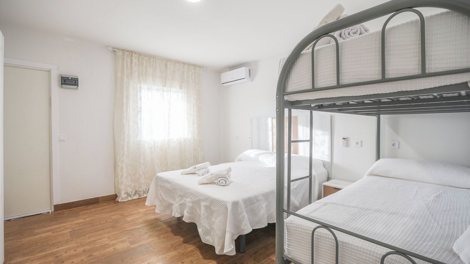 Een kamer met twee bedden, een houten vloer, een raam met dunne gordijnen en een airconditioning.