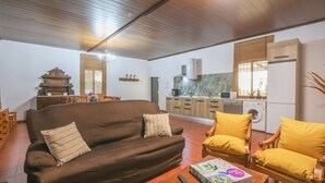 Ein Wohnzimmer mit einer Couch, Sesseln, einem Esstisch und einer Küchenzeile mit Schränken und Geräten.