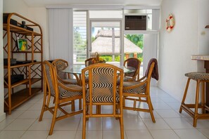 A dining area with wicker chairs, a glass table, a bookshelf, and a view of a thatched-roof structure through a sliding glass door.