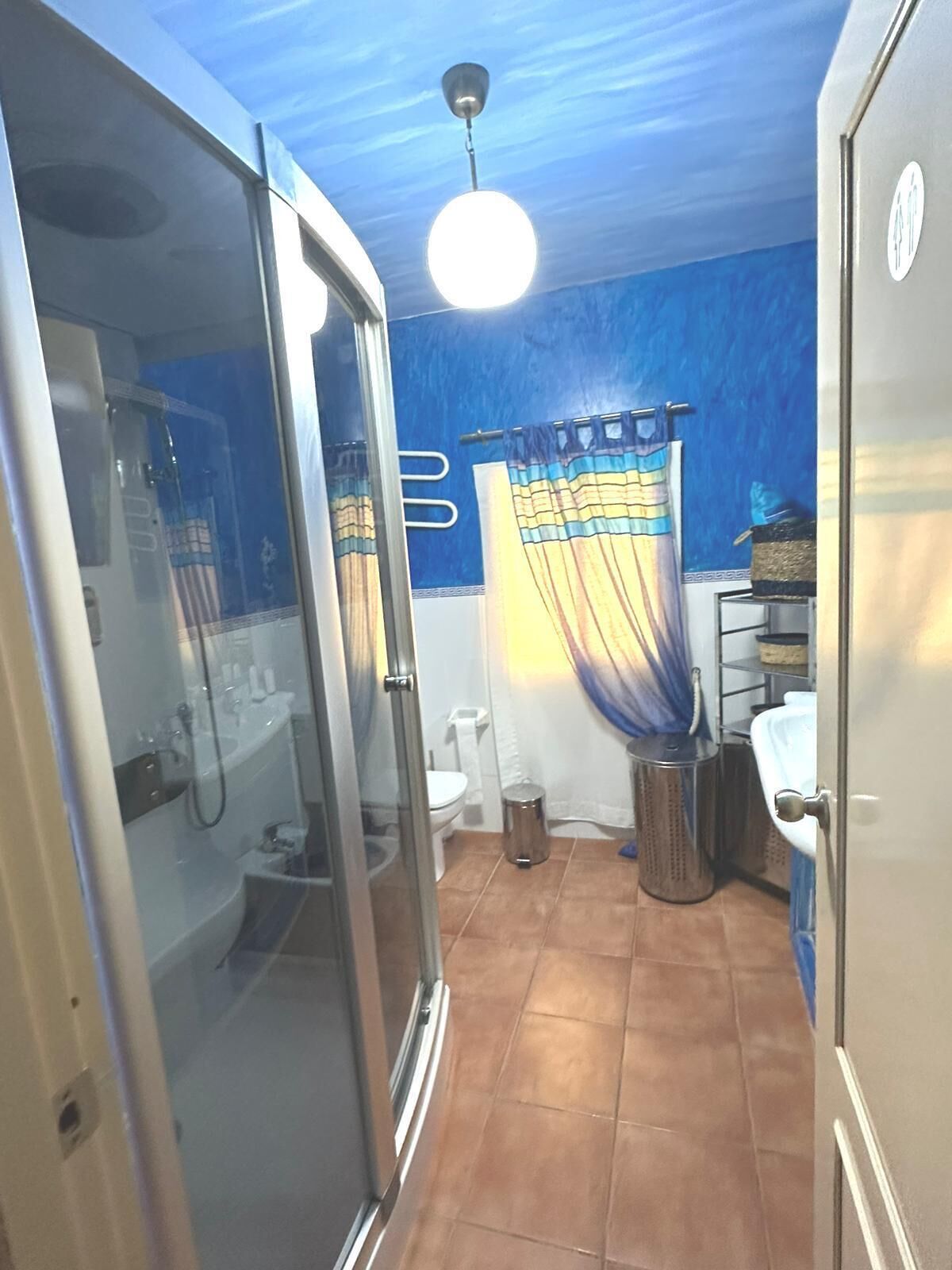 Une salle de bain avec une douche à parois de verre, des toilettes blanches, une poubelle et un luminaire au plafond.