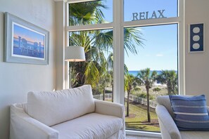 A room with a white sofa, a lamp, a framed picture, and a large window with a 'RELAX' sign. - Luxury Beachfront Getaway 3BR Sleeps 10 Pool+ Beach Access (Destin)