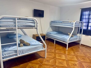 Ein Zimmer mit zwei Etagenbetten, einem an der Wand befestigten Fernseher, einem kleinen Nachttisch aus Holz und einem Fenster mit Vorhängen.