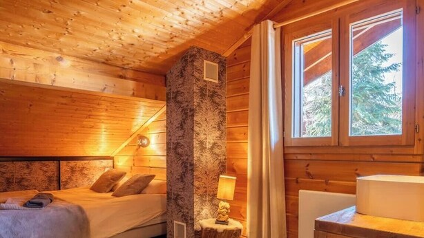 Ein gemütliches Cabin-Zimmer mit Bett, Steinpfeiler, kleinem Tisch mit Lampe und einem Fenster mit Vorhängen.