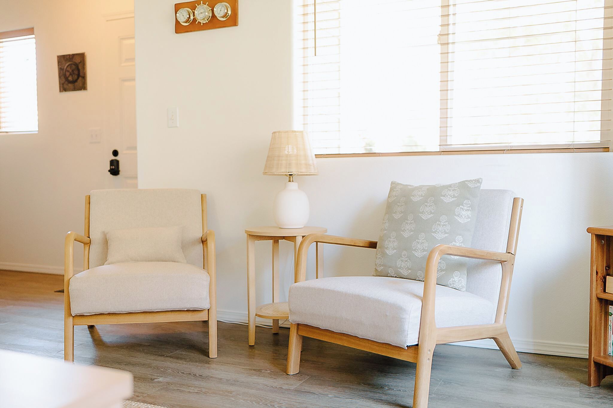 Una habitación con dos sillas de colores claros, una mesita con una lámpara y una estantería.