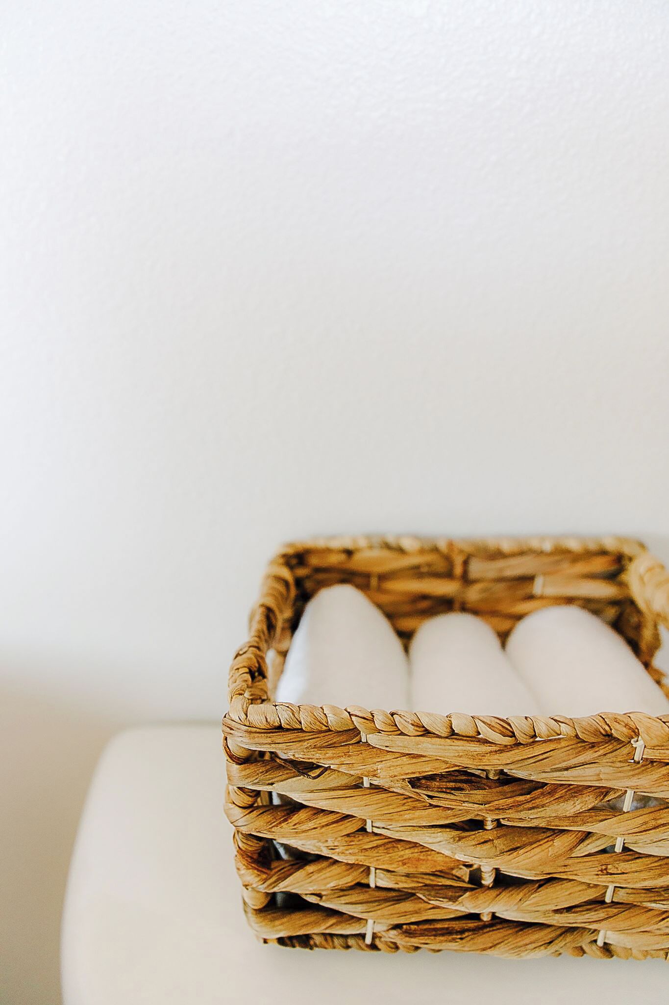 A wicker basket with cylindrical objects, possibly candles, on a white surface against a plain wall.