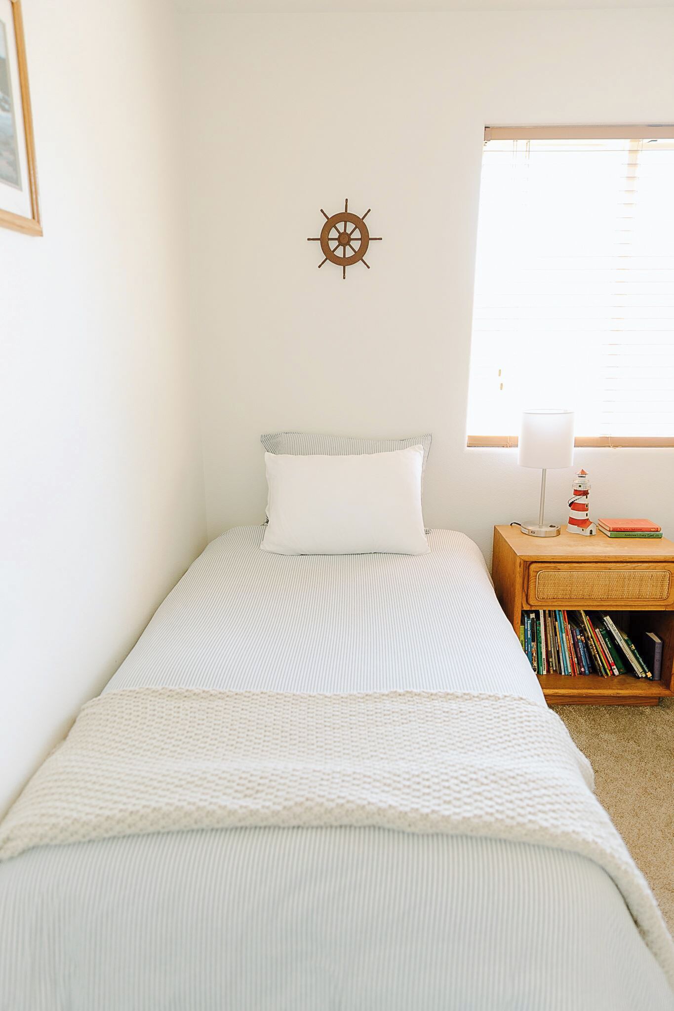 A small, neatly made bed with a striped bedspread, a white pillow, a wall-mounted compass, a bedside lamp, and a small bookshelf with books.