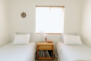 A bedroom with two single beds, a nightstand with a lamp and books, a window with blinds, and a wall-mounted compass. - The Lighthouse - Entire home in Moscow, Idaho (Moscow)