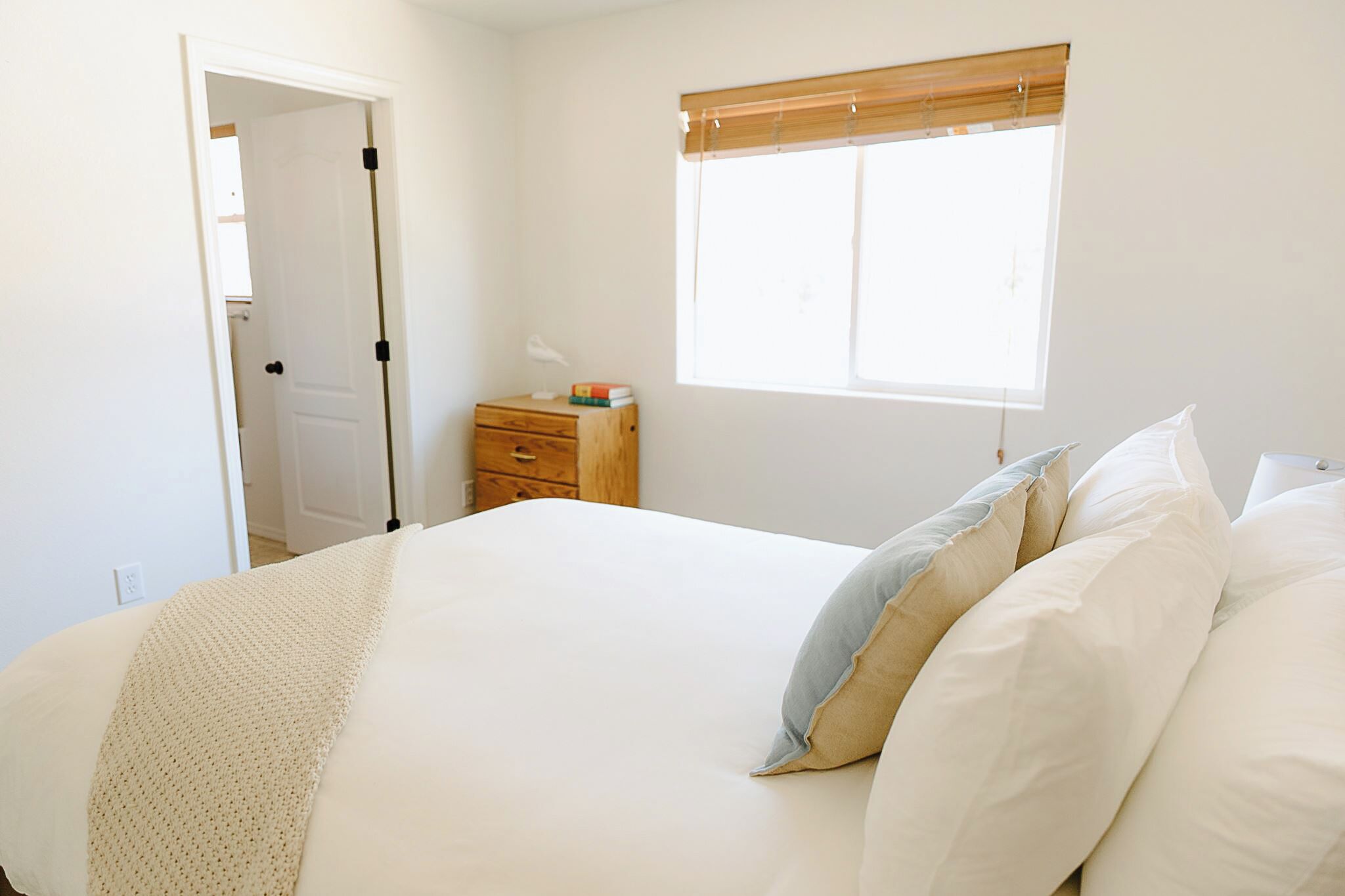 A bedroom with a bed, a nightstand, a window with blinds, and a door.