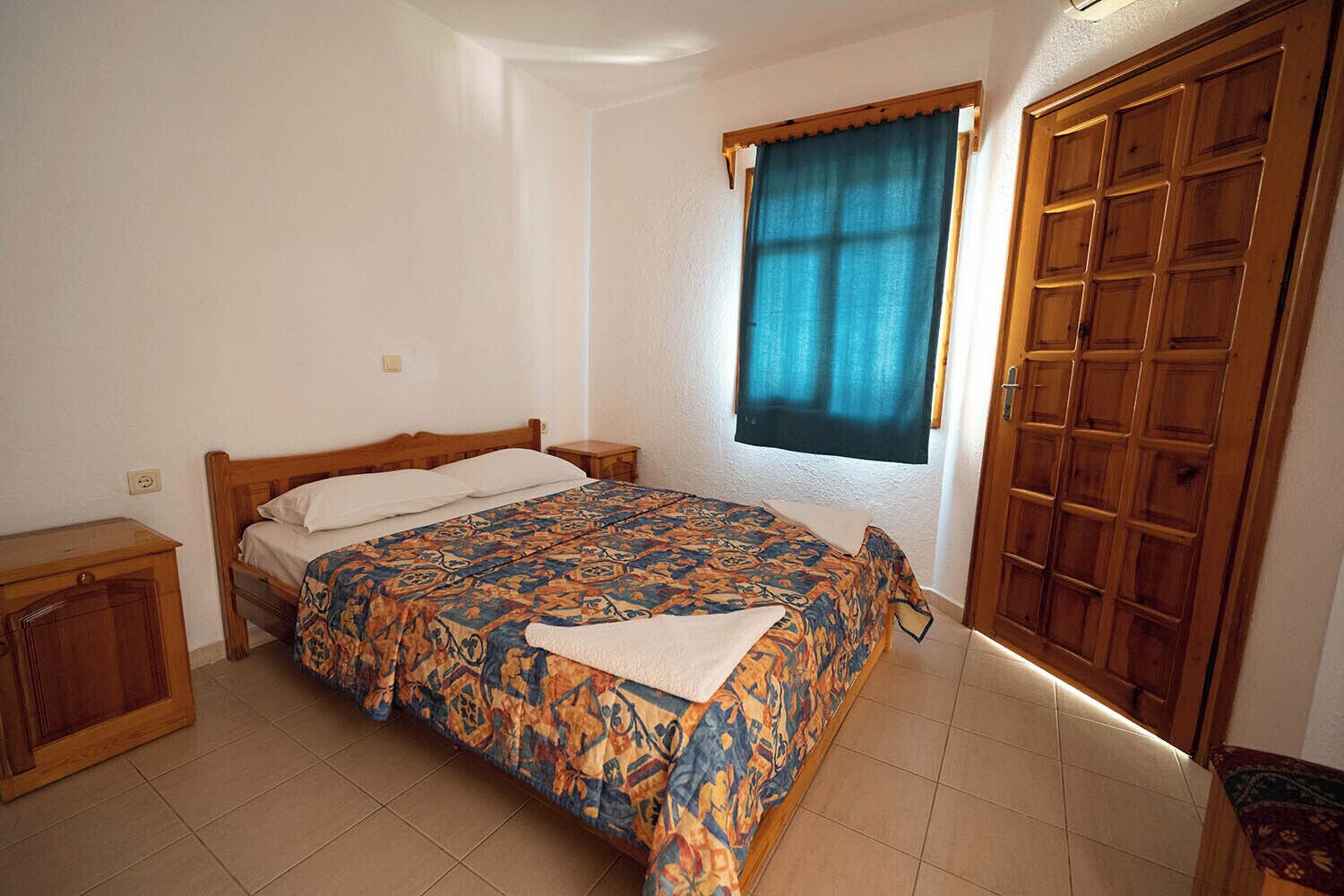 Un dormitorio con cama, armario de madera, ventana con cortina y mesita de noche.