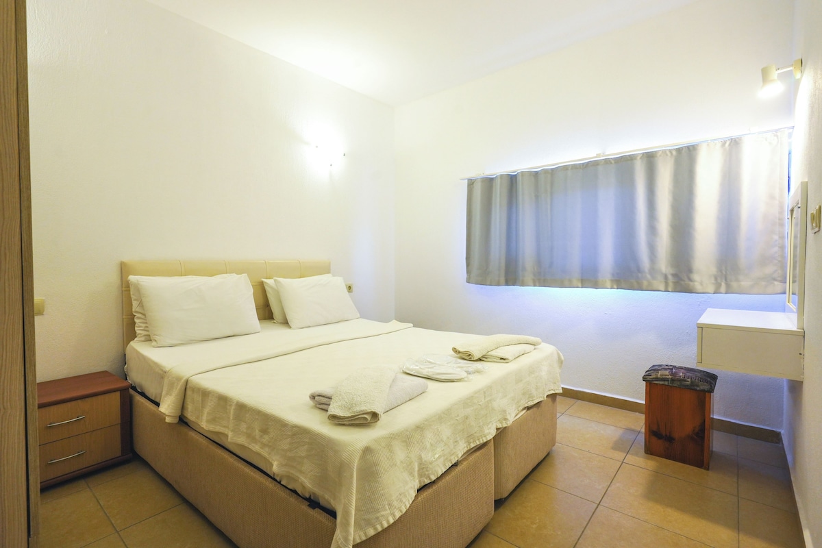 A single-bed room with a wooden headboard, a bedside table, a small stool, and a window with curtains.