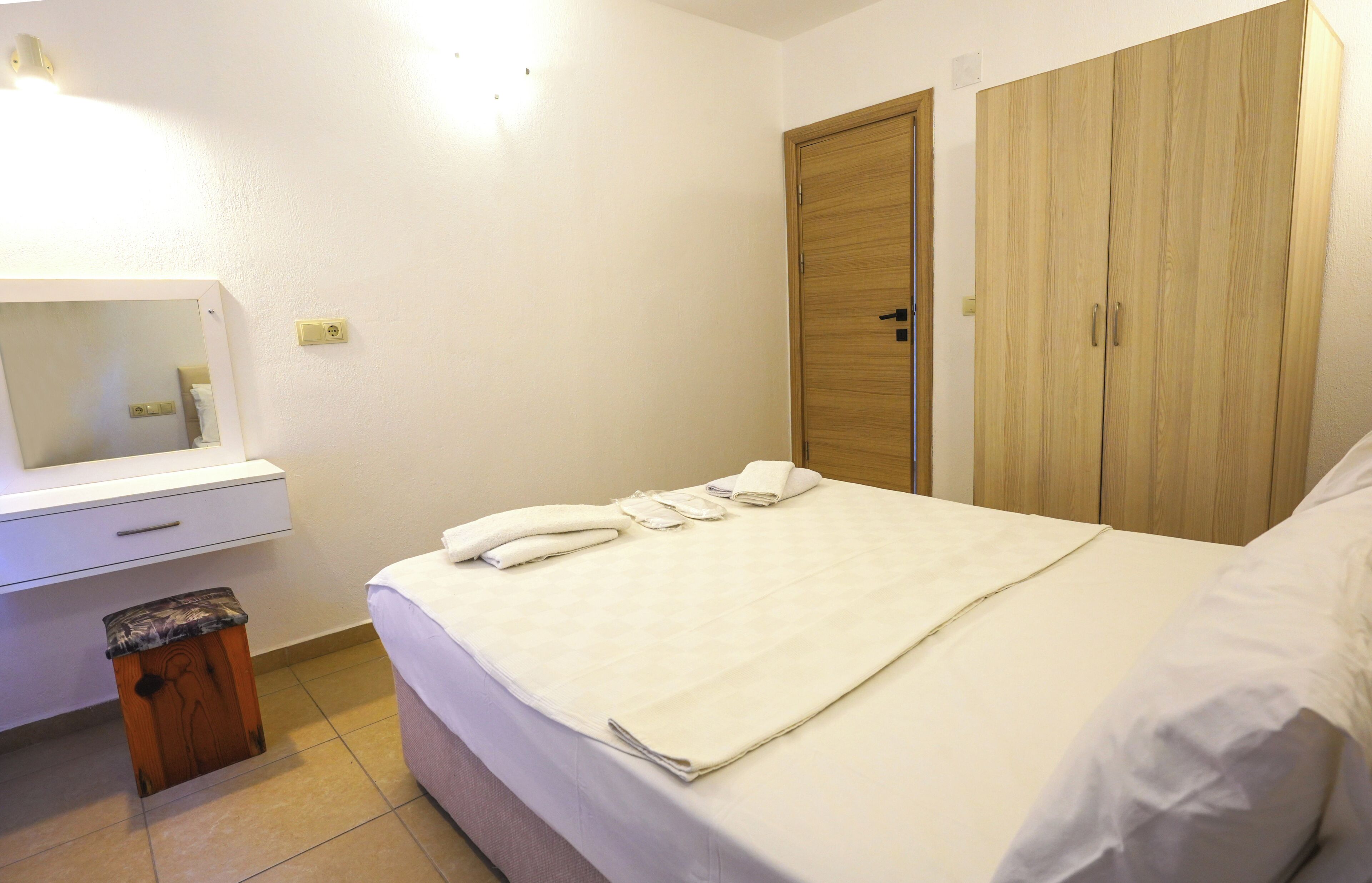 A neatly made bed with a white blanket and pillows, a wooden wardrobe, a white bedside table with a mirror, and a small wooden stool.
