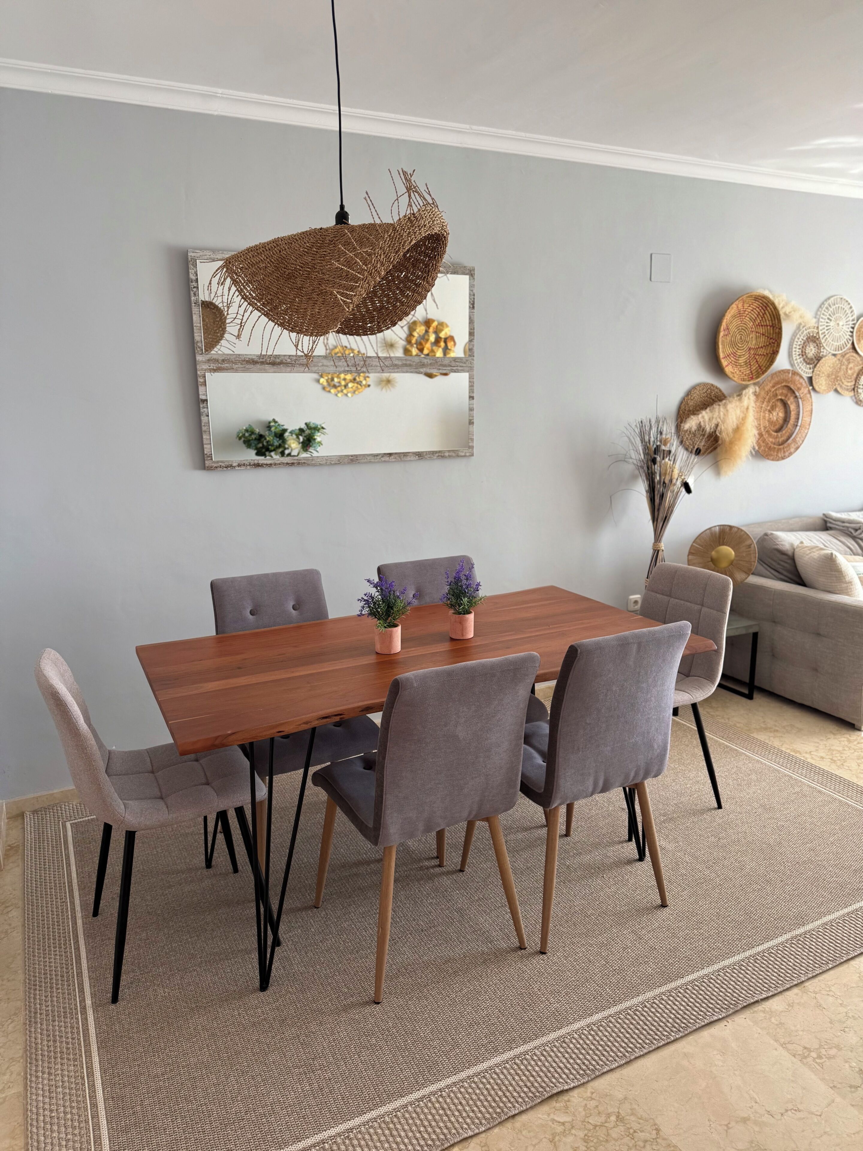 Un espace repas comprenant une table en bois, des chaises à revêtement gris et un luminaire suspendu en fibres tressées.