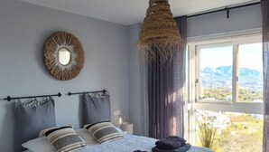 A bedroom with a bed, a hanging light fixture, a window with a view, and a wall-mounted mirror. - Penthouse 2 bed 2 bath Marbella. Breathtaking sea & mountain views. (Ojén)