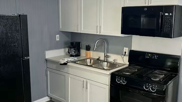 Cocina compacta con gabinetes blancos, un refrigerador negro, microondas, lavamanos y estufa.
