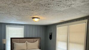 Un dormitorio con cama, mesita de noche, lámpara, luz de techo y ventanas amplias con persianas.
