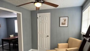Una habitación con ventilador de techo, una puerta, un cuadro enmarcado en la pared, un sillón color beige y una mesa de comedor con sillas.