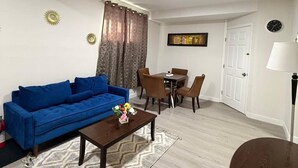 Uma sala de estar com sofá azul, mesa de centro, área de jantar com cadeiras, relógio na parede e uma porta.