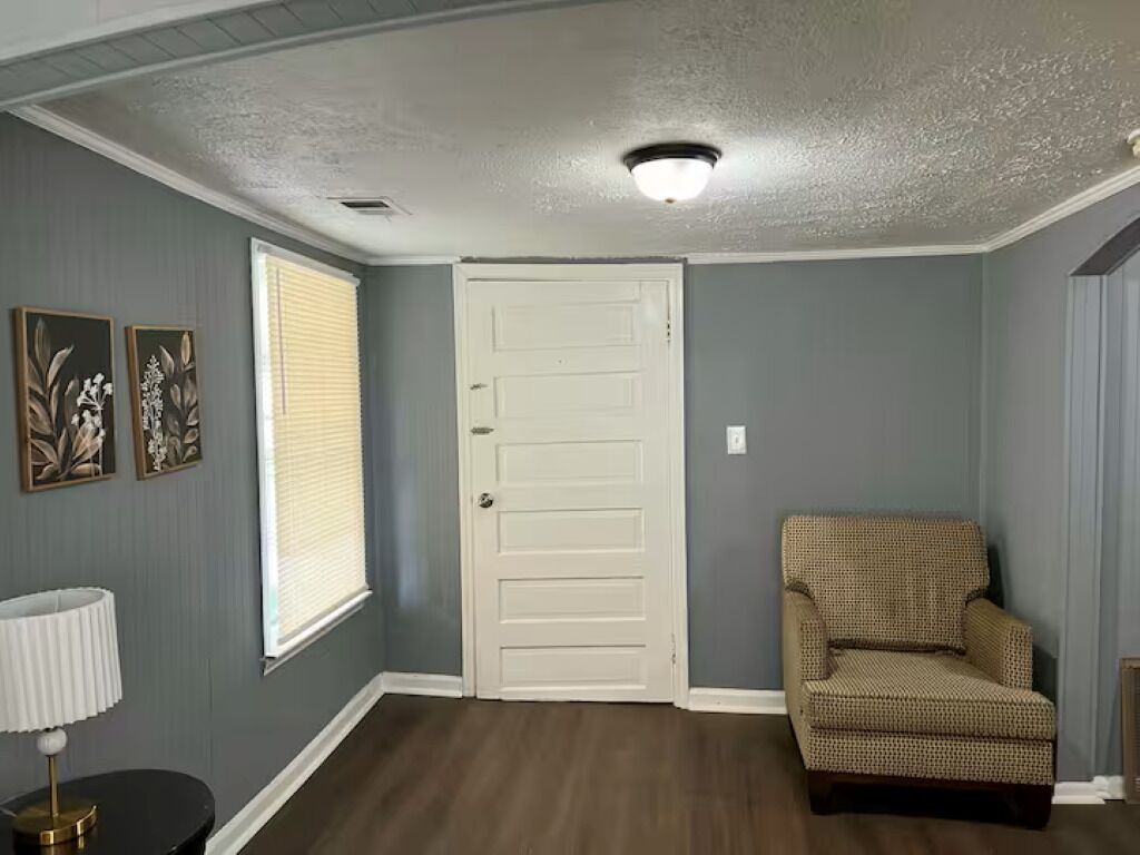 A room with a door, a patterned armchair, a lamp, and framed artwork on the wall.