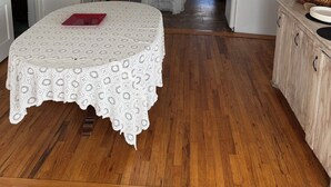 Een keuken met houten vloeren, witte kastjes, een eettafel met een kantone tafellaken en een koelkast.