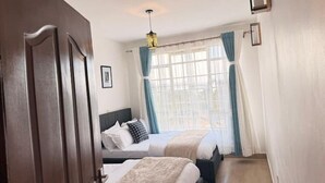 A hotel room with two beds, a large window with curtains, a hanging light fixture, and a wooden door. - Executive 2 bdrm Apartment Meru, Mt. Kenya region (Meru County)