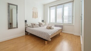 Ein Schlafzimmer mit Bett, großem Fenster, Wandspiegel und Nachttisch mit Lampe.