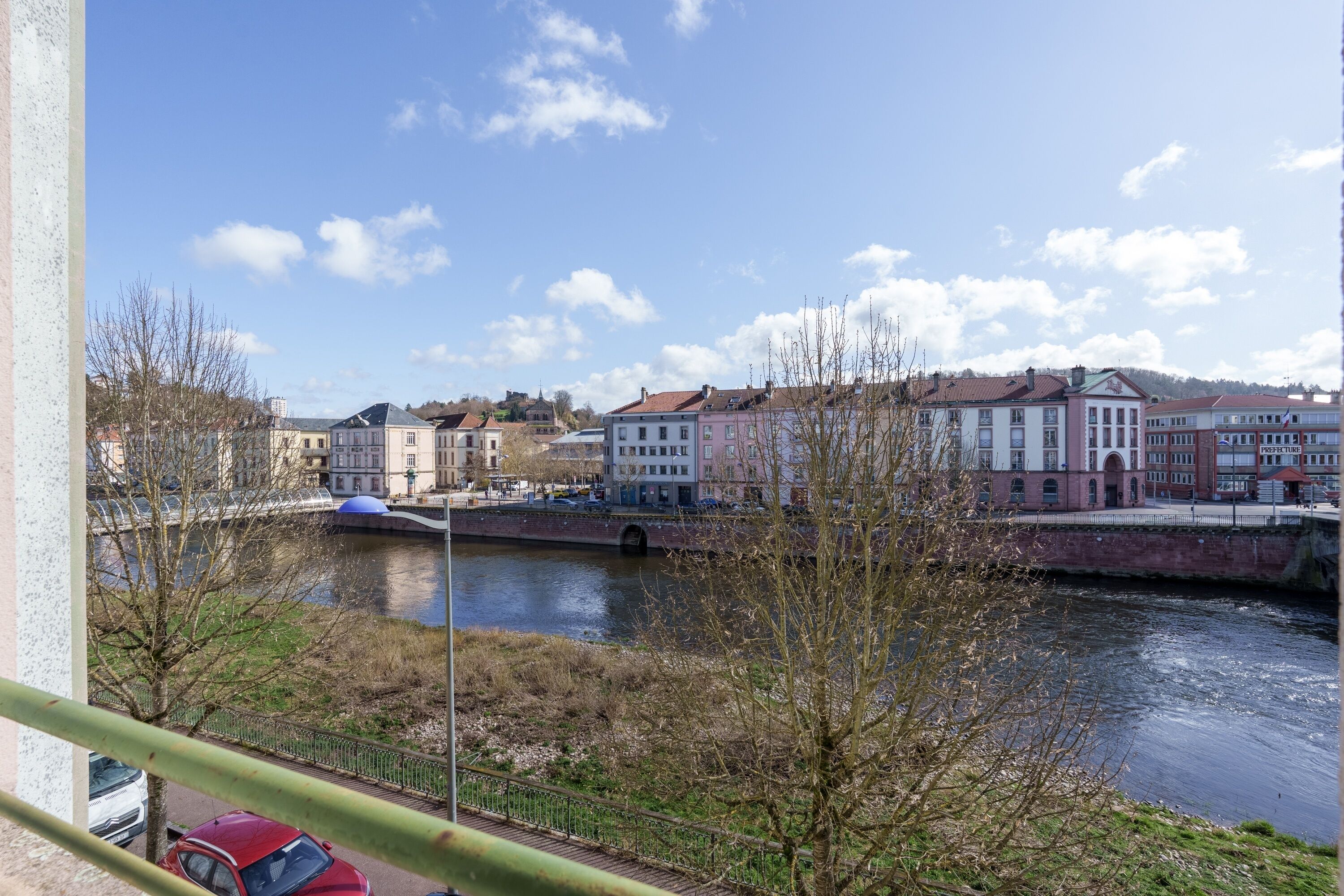 A river with buildings on the opposite bank, a bridge, and a red car parked near the river.
