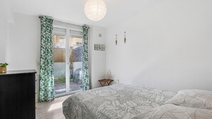 Un dormitorio con una cama, una mesita de noche de madera, una planta en maceta y vistas a través de un gran ventanal corredero.