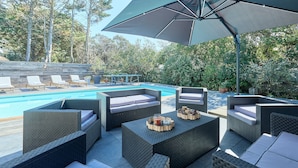 Zona junto a la piscina con muebles de mimbre, una sombrilla grande y una alberca.