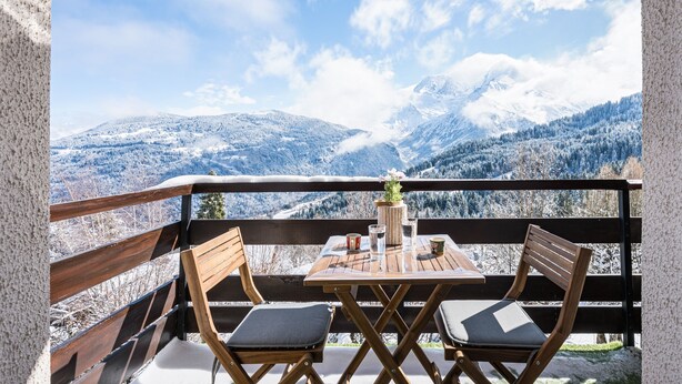 Een balkon met een houten tafel en twee stoelen, met uitzicht op een met sneeuw bedekte berglandschap.