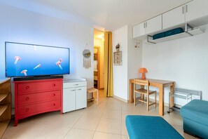 Ein gemütlicher Wohnraum mit Fernseher, einer roten Kommode, einem kleinen Tisch mit Lampe, einem blauen Sofa und einem Kleiderschrank.