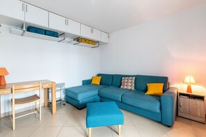 Ein Wohnzimmer mit einem Ecksofa, einem Holztisch, einem Sessel, einem kleinen Hocker, einem Beistelltisch mit Lampe und einbauartigen Regalen über dem Sofa.