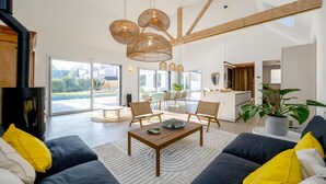 A spacious living area with a large window, a dining table, and a kitchen in the background.