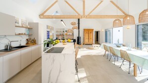 A modern kitchen with a marble island, wooden beams, pendant lights, and a dining area with a long table and chairs.