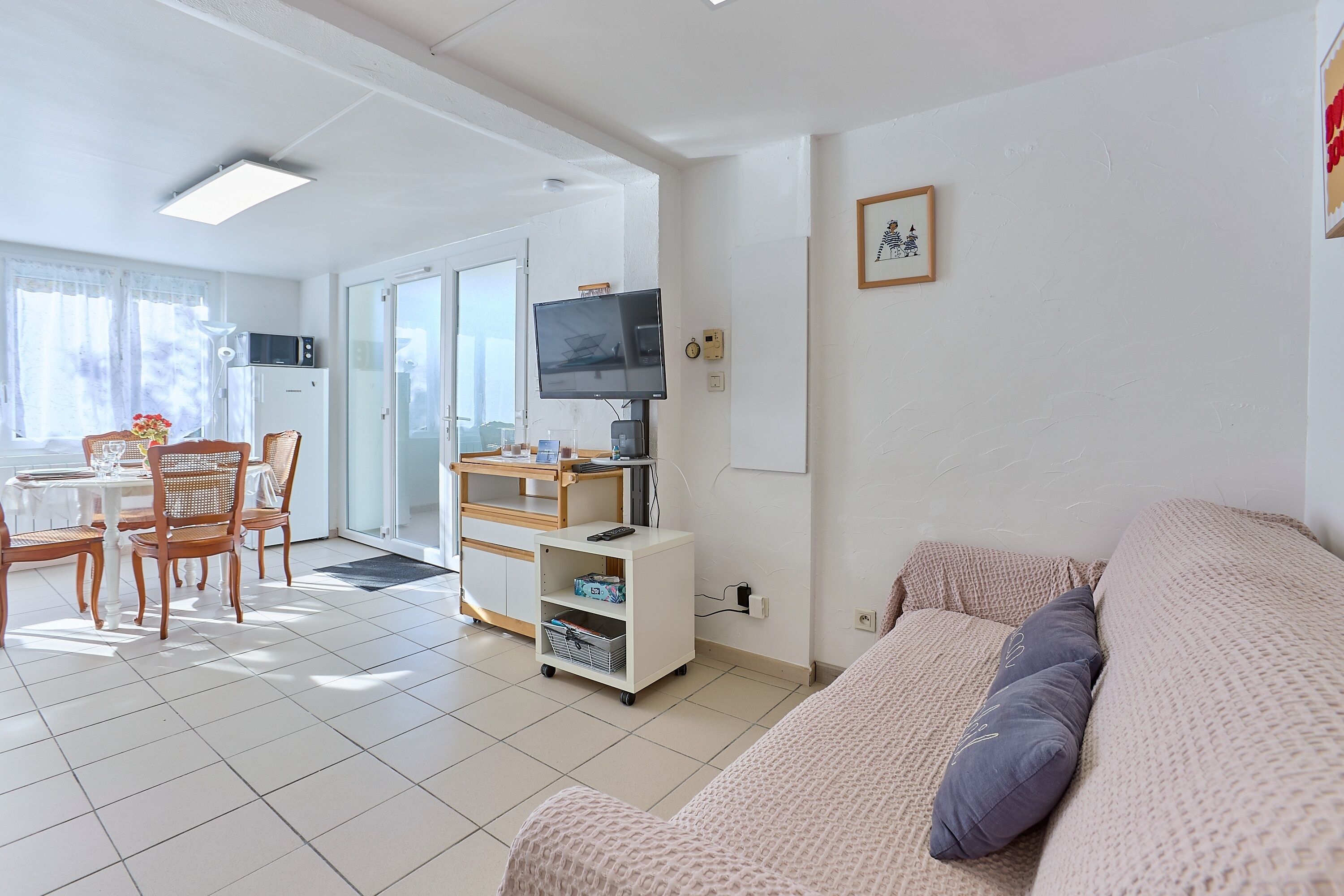 A living room with a sofa, a TV, a dining table with chairs, a kitchen area, and a sliding glass door leading to a balcony.
