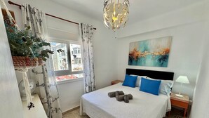 A bedroom with a bed, a nightstand, a lamp, a fan, a window with curtains, a hanging plant, and a painting on the wall. - Poppy of SteraM Flats Torremolinos (Torremolinos)