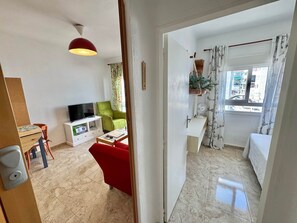 Ein Wohnbereich mit Fernseher, grünem Sessel, rotem Sofa und einem kleinen Tisch mit Ventilator.