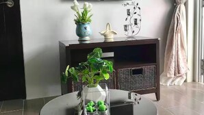 Une pièce avec un téléviseur fixé au mur, un meuble en bois foncé agrémenté d’objets décoratifs, un vase en verre contenant des plantes et un fauteuil en osier.