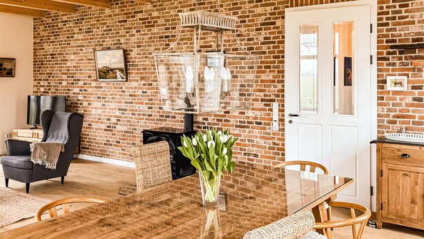 A room with a brick wall, a dining table with chairs, a television, a fireplace, and a door. - Cozy home in Detern with WiFi (Detern)